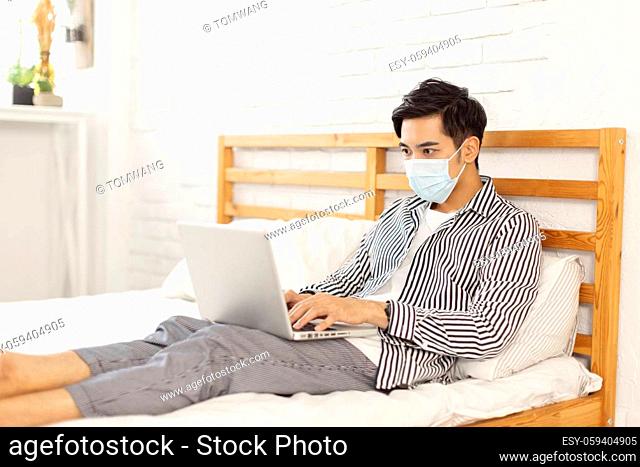 young man wearing a medical mask , lying in bed, using a laptop to work