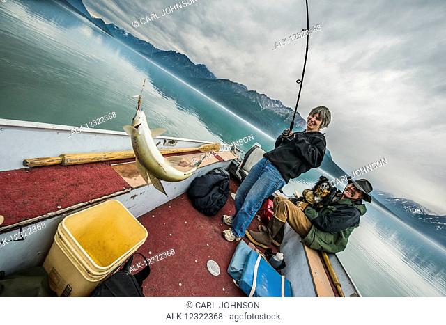 A woman catches a greyling from a small boat while her husband and dog watch, Lake Clark National Park & Preserve, Southcentral Alaska, USA