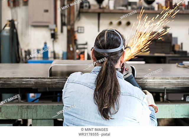 Rear view of woman wearing safety glasses and dust mask standing in metal workshop, using power grinder, sparks flying