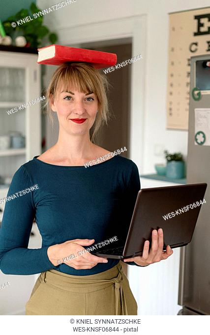 Portrait of smiling woman standing with laptop in the kitchen balancing book on her head