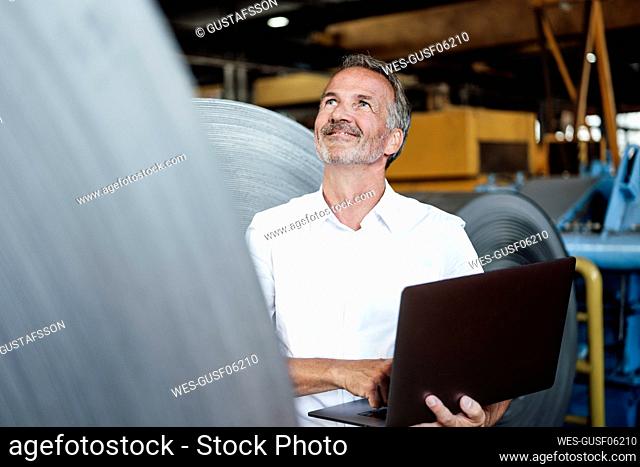 Smiling male inspector with laptop checking metal sheet roll in steel mill