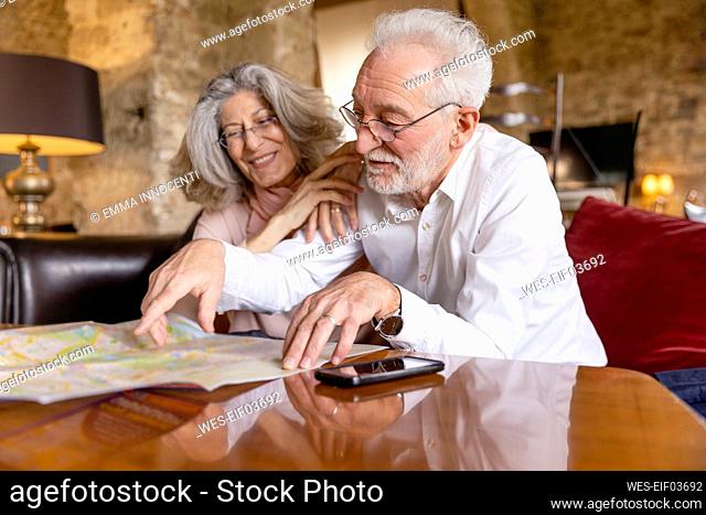 Smiling senior man pointing at map sitting by woman on sofa at boutique hotel