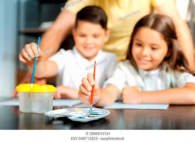 Young artists. The focus being on the hands of lovely smiling girl choosing a necessary color from the palette and her brother washing his brush in a water...