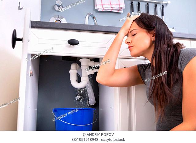 Worried Woman Collecting Water Leaking From Sink In Blue Bucket