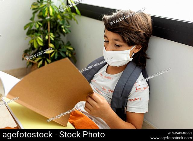 Close-up of boy wearing mask reading book while sitting against wall in school