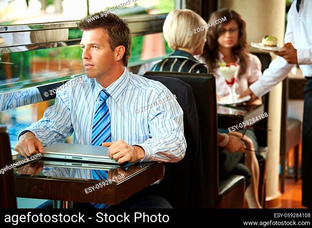 Businessman sitting at coffe table in cafe, waiter serving sweets to young women in the background