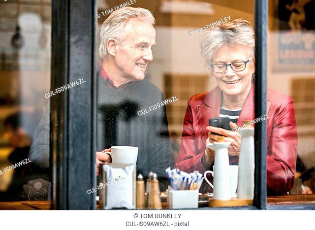 Senior couple in cafe window seat drinking coffee and texting on smartphone