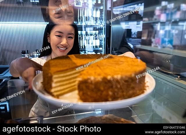 Cheerful cafe worker taking a serving platter with a tasty dessert from the glass pastry display case shelf