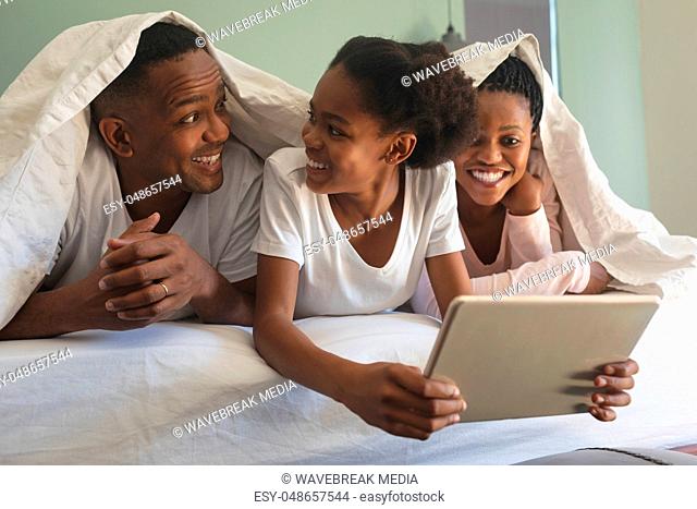 Happy African American family under blanket and using digital tablet on bed in bedroom