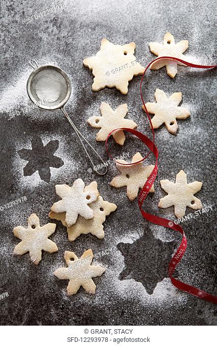 A number of snowflake shaped biscuits in the process of being dusted with icing sugar on a grey slate background mini sifter and red Noel ribbon