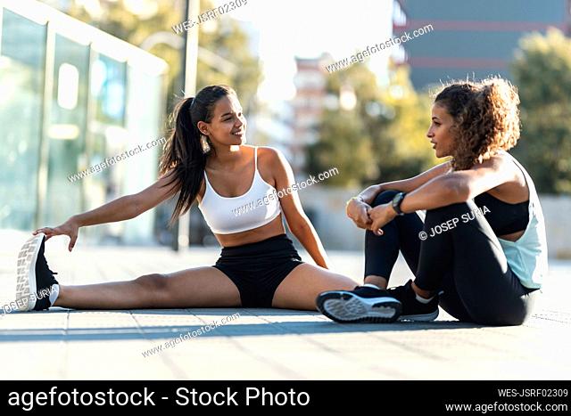 Happy teenage girl exercising and talking to friend sitting on footpath