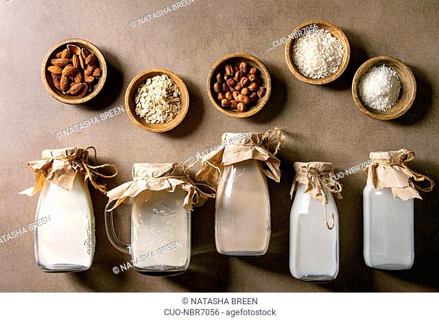 Variety of non-dairy vegan lactose free nuts and grain milk almond, hazelnut, coconut, rice, oat in glass bottles in row with ingredients above over brown...