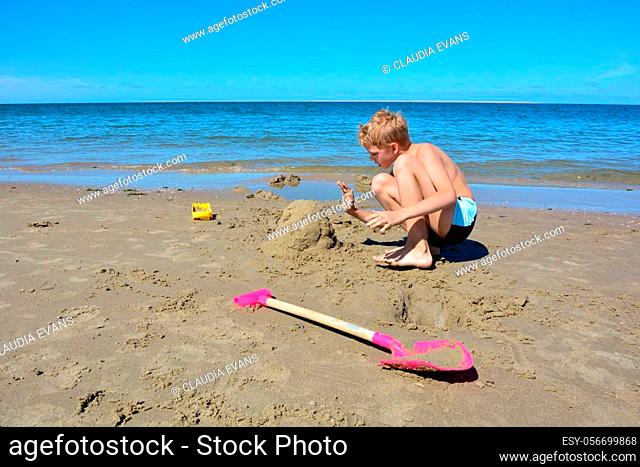 A little blond boy with swimming trunks builds a sand castle in the sand on the beach in front of the sea, with a blue sky