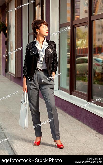 Happy young fashion woman in leather jacket with handbag next to mall window