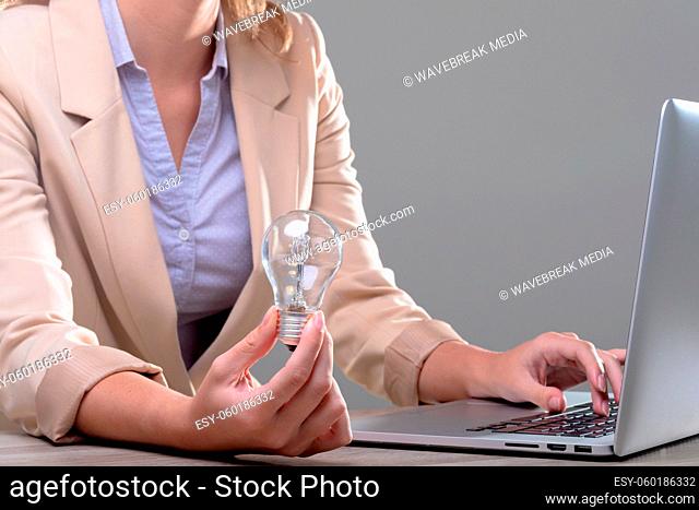 Midsection of caucasian businesswoman holding light bulb using laptop, isolated on grey background