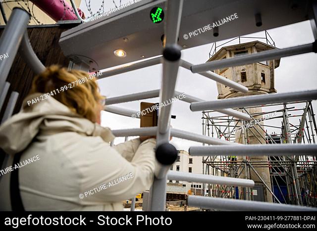11 April 2023, Berlin: A woman takes a photo through a revolving door in a fence of a GDR watchtower of the type BT-6 near Potsdamer Platz in...