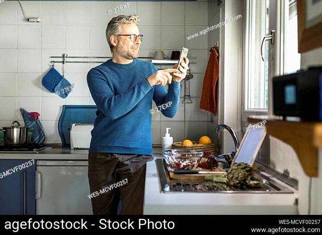 Smiling mature man standing in his kitchen taking selfie with smartphone
