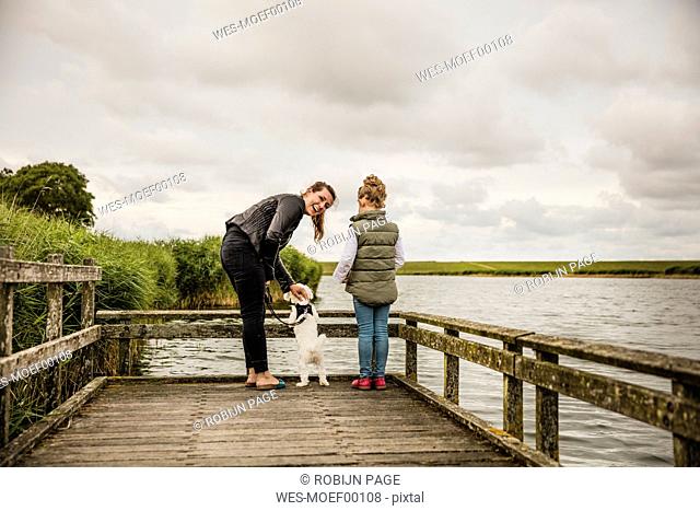 Mother and daughter standing on jetty at a lake with dog