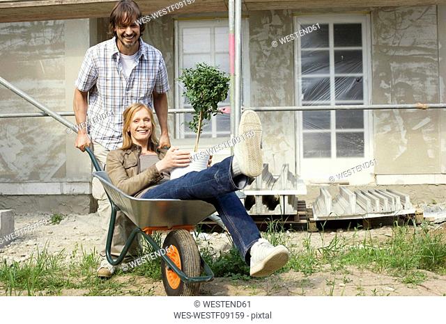 Young couple at construction site, man pushing woman in wheelbarrow