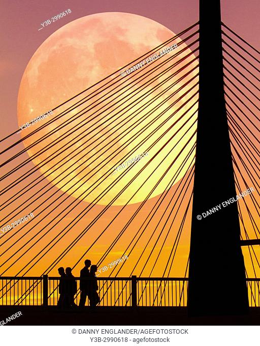 Silhouettes of people at sunset, walking near the Zakim Bridge in Boston, Massachusetts, as the full moon rises