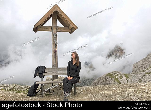 jeune femme assise sur un banc sous un crucifix sur le chemin reliant les refuges Locatelli (Dreizinnenhutte) et Pian di Cengia (Bullelejochhutte)
