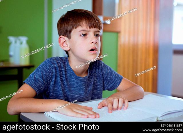 Blind caucasian schoolboy sitting at desk in classroom reading braille book with fingers