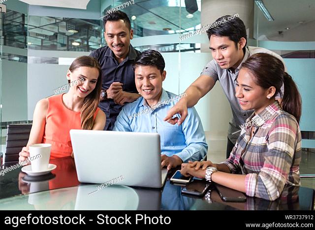 Multi-ethnic team of five cheerful employees smiling while watching a funny video or presentation on laptop in the meeting room of a multinational company