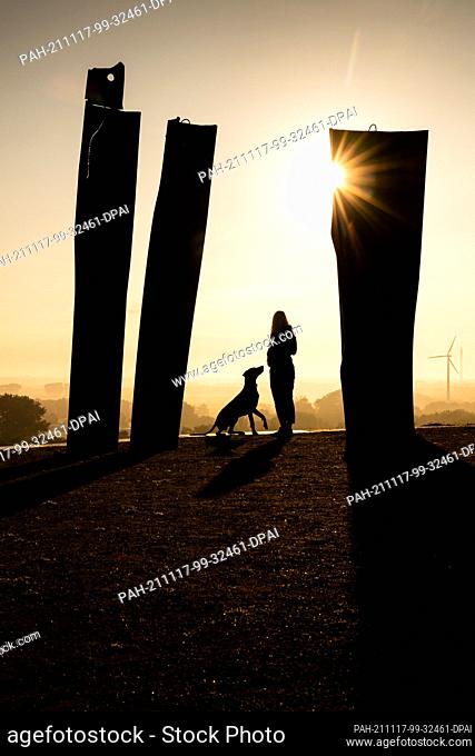 14 September 2021, Bremen: A woman stands with her dog at sunrise at the Metalhenge viewpoint on the Blockland Landfill. Photo: Sina Schuldt/dpa
