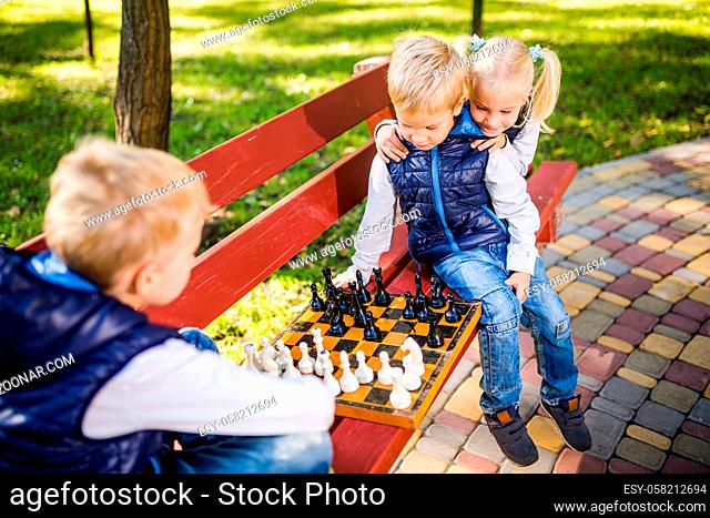 Caucasian children play chess on wooden chessboard in park bench. Brothers and sister big friendly active mental family spend time on vacation in sunny weather