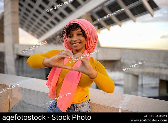 Smiling young woman gesturing heart shape standing by wall