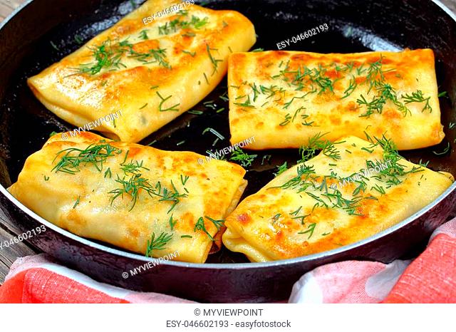 delicious mozzarella stuffed crepes wraps on skillet sprinkled with finely chopped dill, on dark wooden board, view from above, close-up