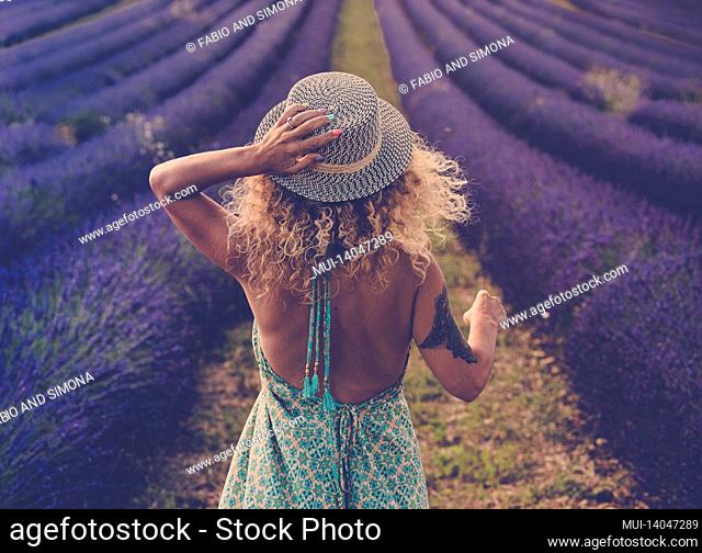 back view of pretty babe in boho elegant blue dress walk in lavender fields wearing travel style hat and blonde curly nice long hair - concept or free woman in...