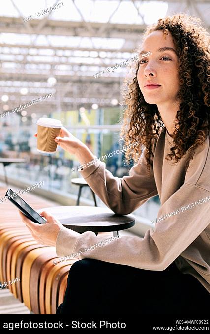 Thoughtful woman sitting with mobile phone drinking coffee at train station