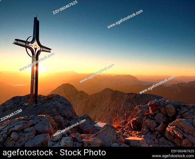Cross raised at mountain summit in Alps. Sharp peak, daybreak Sun in sky. Steel crucifix in memory of victims of mountains. Vivid photo