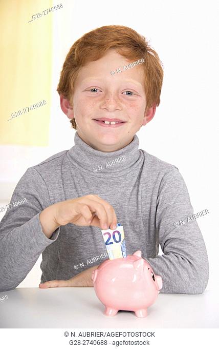 Little boy with red hair, happy putting a 20 euros banknote into the slot of his pink piggy bank