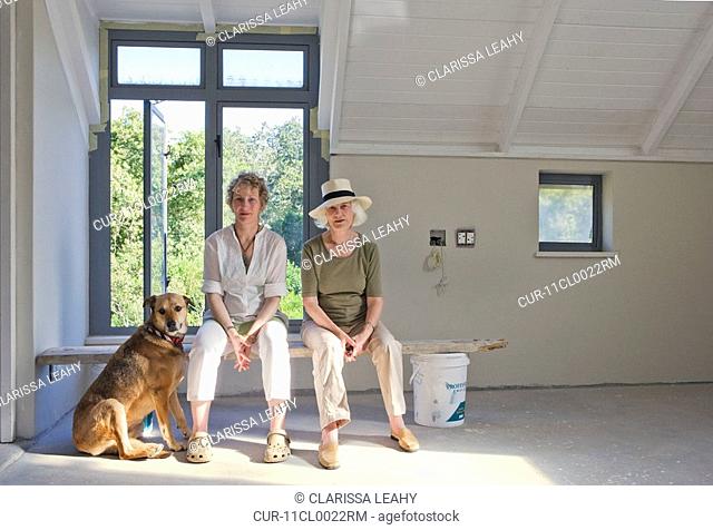 Older mother and daughter sitting on plank with dog in newly built house