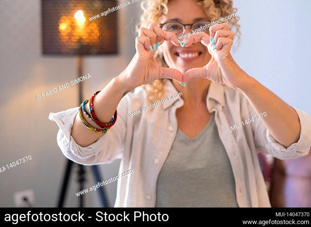 happy beautiful adult portrait of woman doing heart shape with hands in front of the camera with house indoor background - cheerful female people love and enjoy...