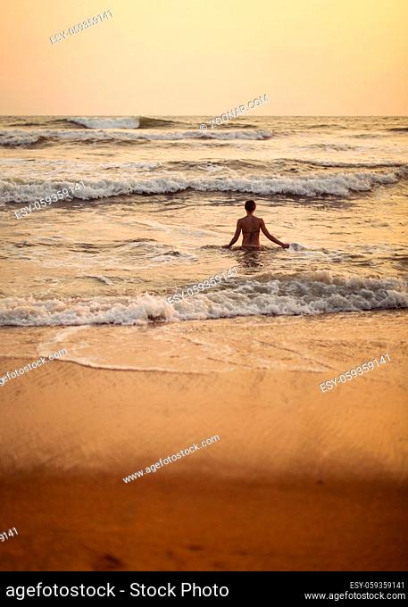 Silhouette of young woman going into the sea with small waves during golden evening light just after the sunset