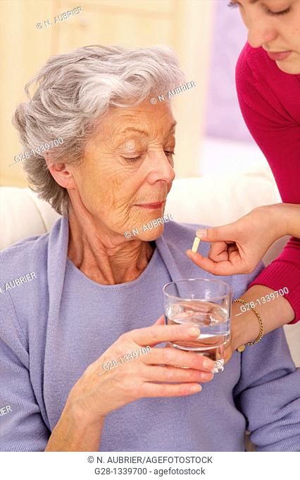 senior woman in a mauve cardigan, holding a glass of water, and young help giving her a whited tablet at home