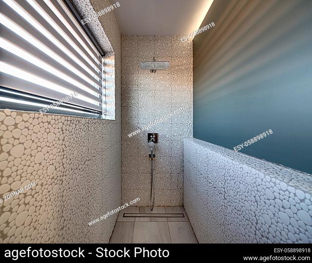 Bathroom in a modern style with decorative tiles and showers on the wall. There is a window with the blinds on the left. Sunlight falls through the blinds