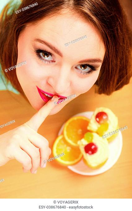 Woman eating delicious gourmet sweet cream cake cupcake and orange showing hand quiet sign gesture. Glutton girl having breakfast in secrecy