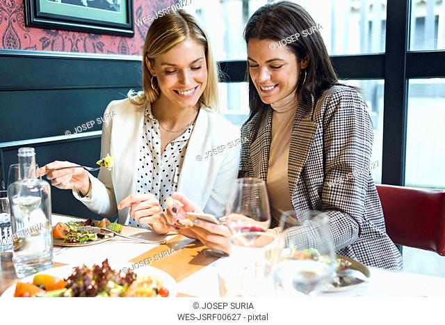 Two happy businesswomen in a restaurant looking at smartphone together