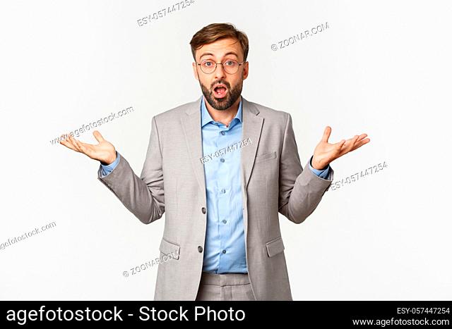 Portrait of surprised businessman with long beard, wearing grey suit and glasses, spread hands sideways and open mouth confused