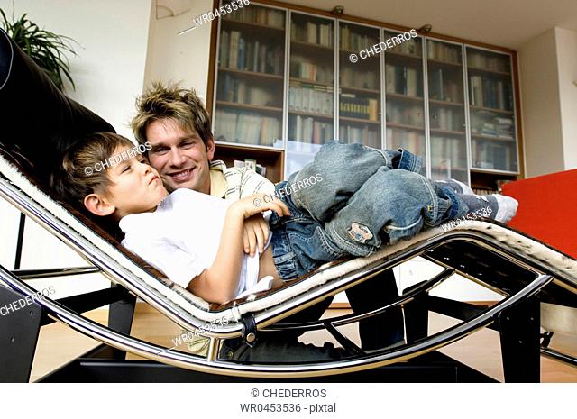 Side profile of a son lying on a chaise longue with his father sitting beside him