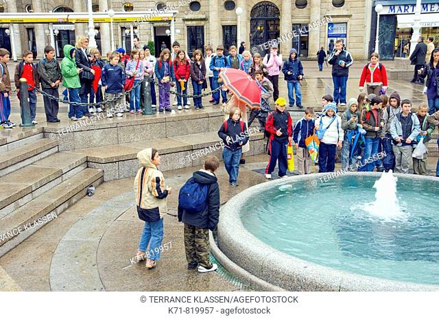 A group of school children with their teacher gather around the Mandusevac Fountain in Ban Jelacic Square on a raining day in Zagreb, Croatia