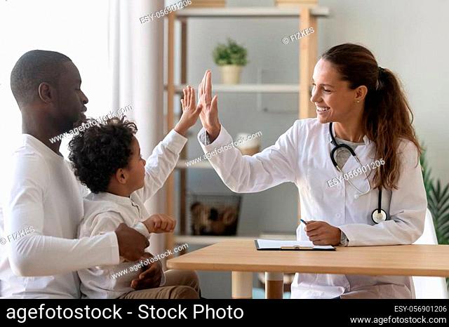 Happy mixed race african cute little child boy give high five to female doctor pediatrician welcome small kid patient and his dad at medical check up...