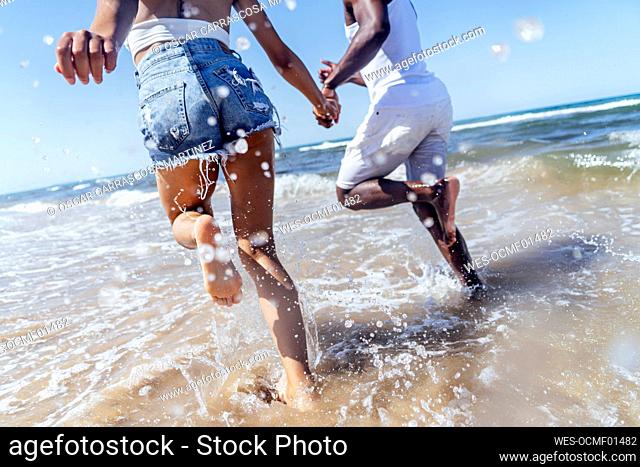 Playful couple holding hands while running in sea against clear sky
