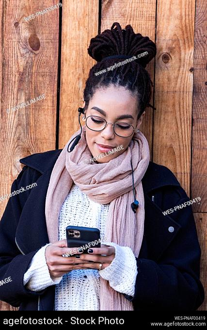 Beautiful young woman using smart phone while leaning against wooden wall