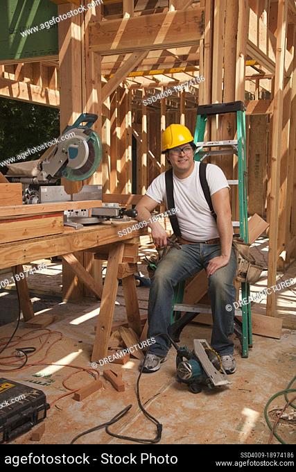 Man at a residential construction site sitting on a ladder and leaning on a work table with a table saw