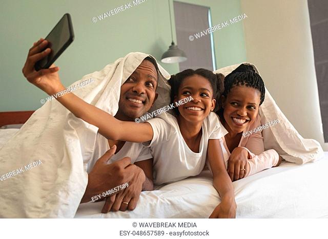 Happy African American family under blanket and taking selfie with mobile phone on bed